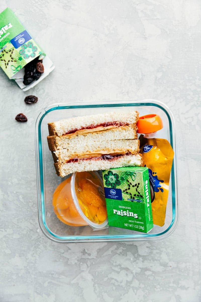 A container filled with a peanut butter and jam sandwich, raisins, mandarin oranges, and apple sauce, all neatly arranged and ready to go.