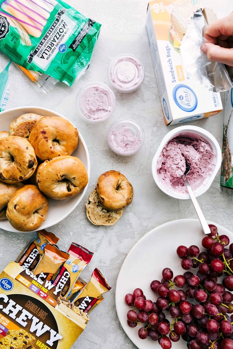 A healthy school lunch, including bagels, string cheese, cream cheese, grapes, and granola bars, arranged for straightforward packing.