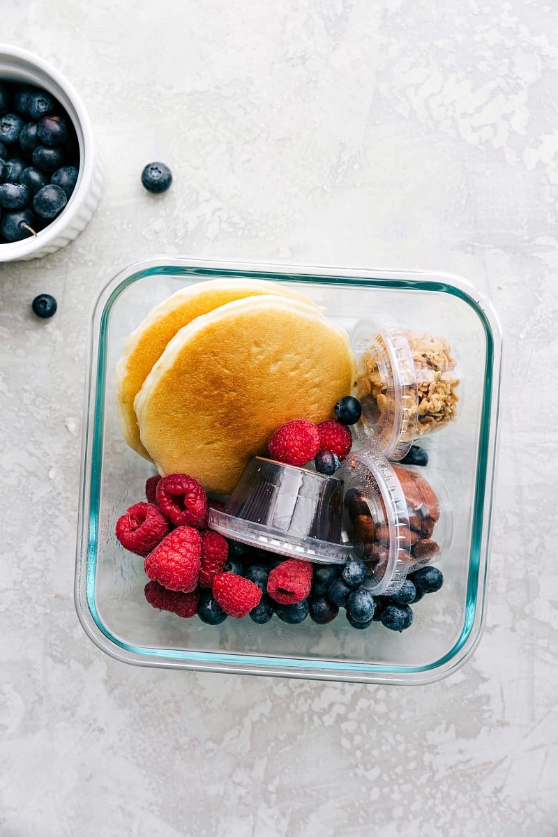Pancakes, syrup, granola, nuts, and fruit packed in containers, ready to go for a wholesome kids school lunch.