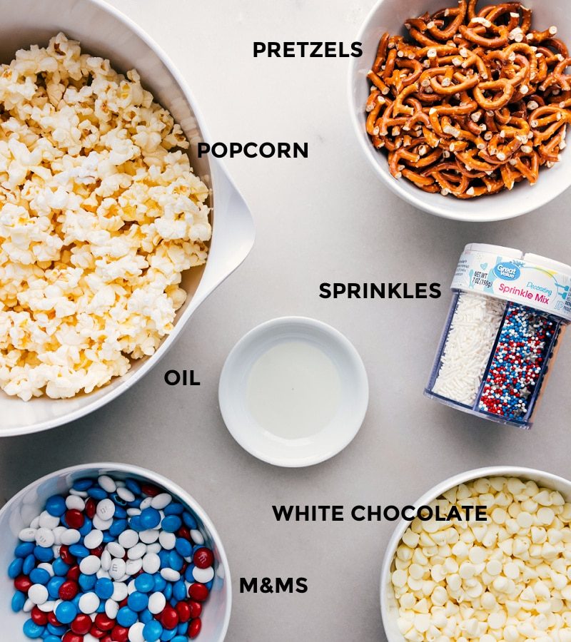 All the ingredients for 4th of July Popcorn laid out, including popcorn, pretzels, sprinkles, white chocolate, and M&Ms, ready for straightforward assembly.