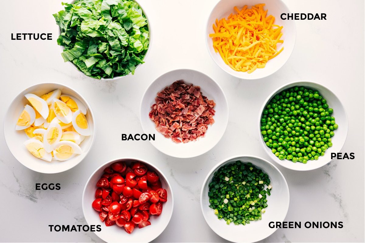 All the ingredients in this recipe prepped out for straightforward assembly including the lettuce, eggs, tomatoes, bacon, green onions, peas, and cheddar.