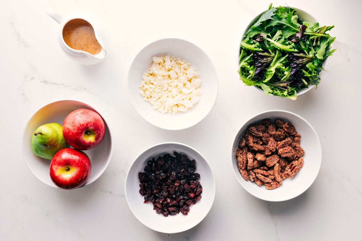 All the ingredients in this recipe prepped out for straightforward assembly including the apples, pear, dried cranberries, pecans, mixed greens, feta, and dressing.