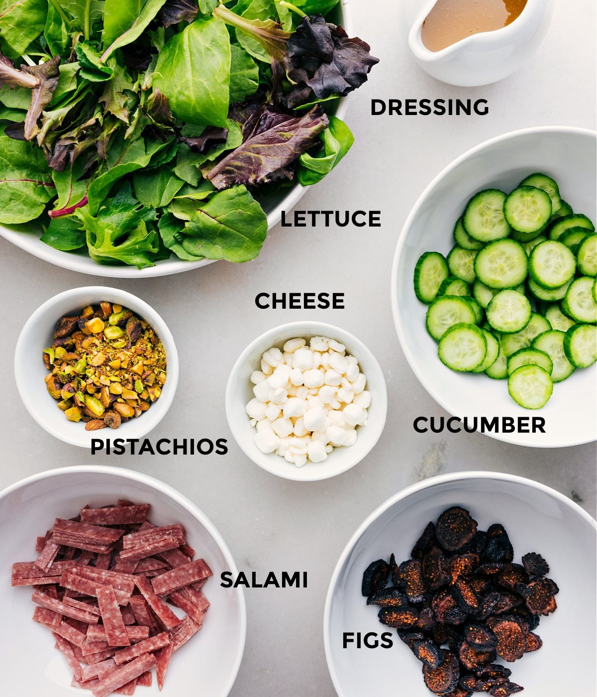 All the ingredients in this recipe—greens, dressing, cucumbers, cheese, pistachios, salami, and figs—are prepped for straightforward assembly.