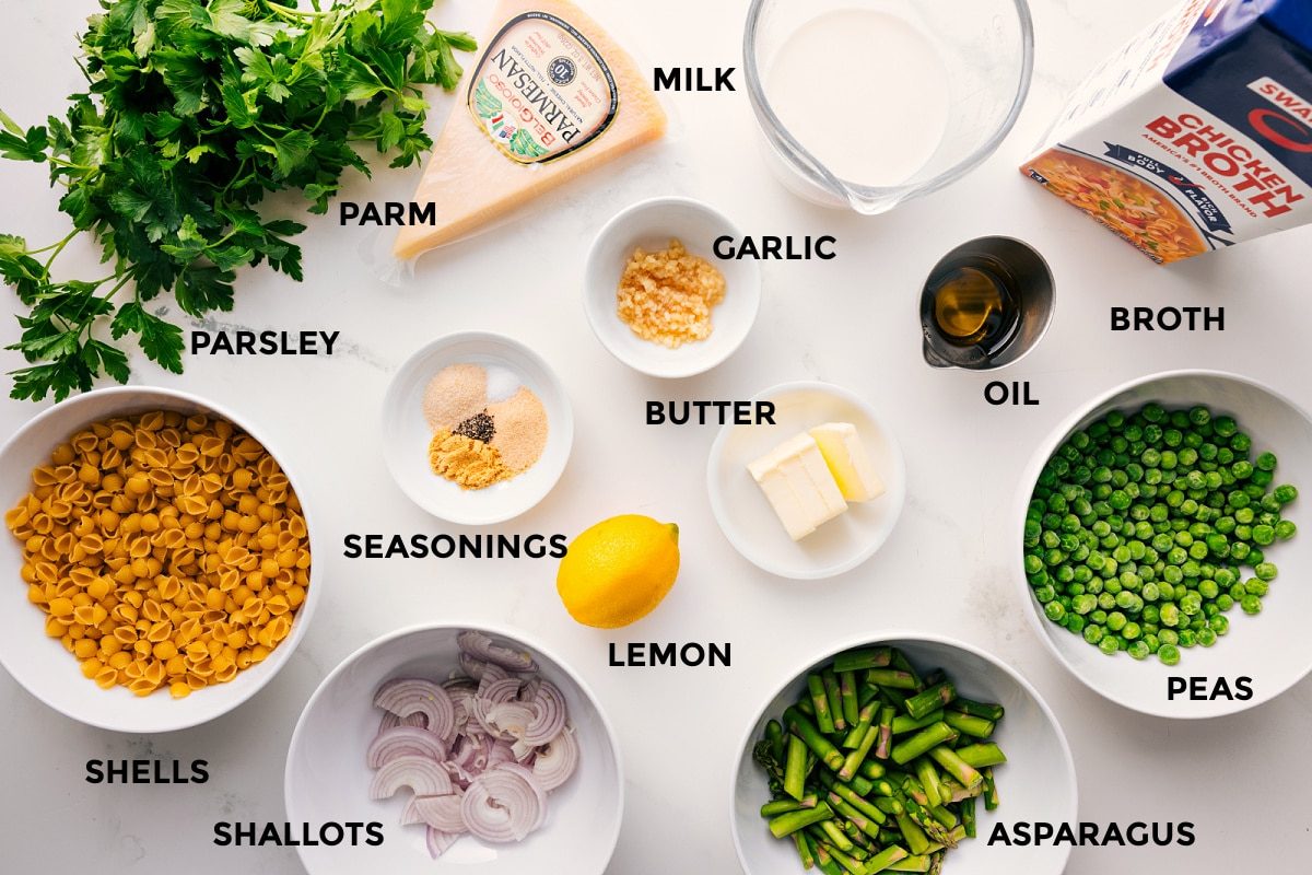 All the ingredients in this recipe prepped out for straightforward assembly including the parsley, shells, shallots, asparagus, peas, lemon, seasonings, butter, garlic, oil, broth, milk, and parmesan.