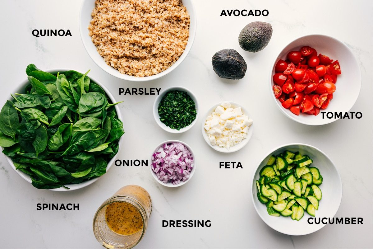 All the ingredients prepped for straightforward assembly, including quinoa, avocado, tomato, feta, cucumber, parsley, onion, dressing, and spinach.
