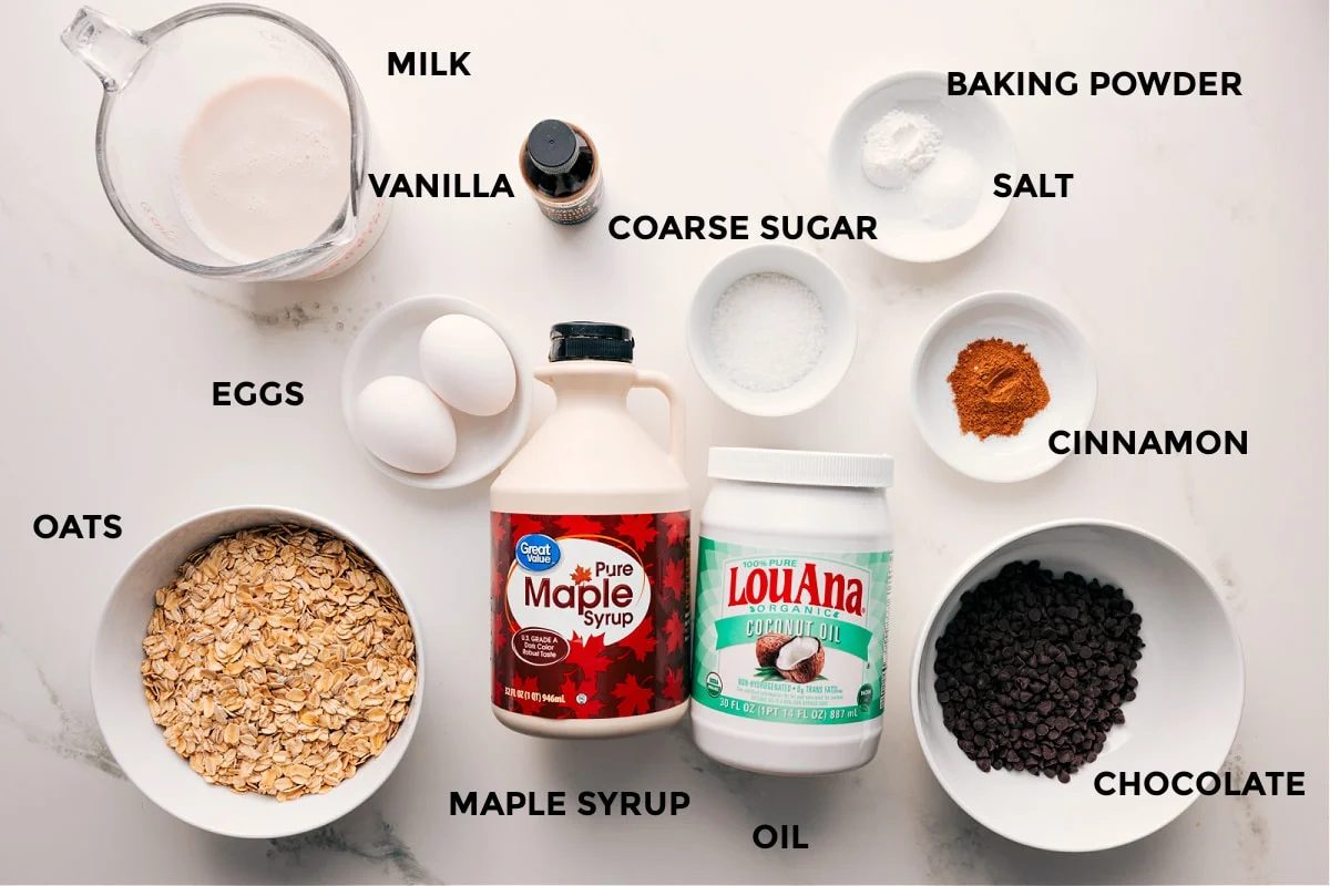 Ingredients in this recipe prepped out for straightforward assembly including milk, vanilla, oats, eggs, maple syrup, oil, chocolate, cinnamon, baking powder, salt, and coarse sugar.