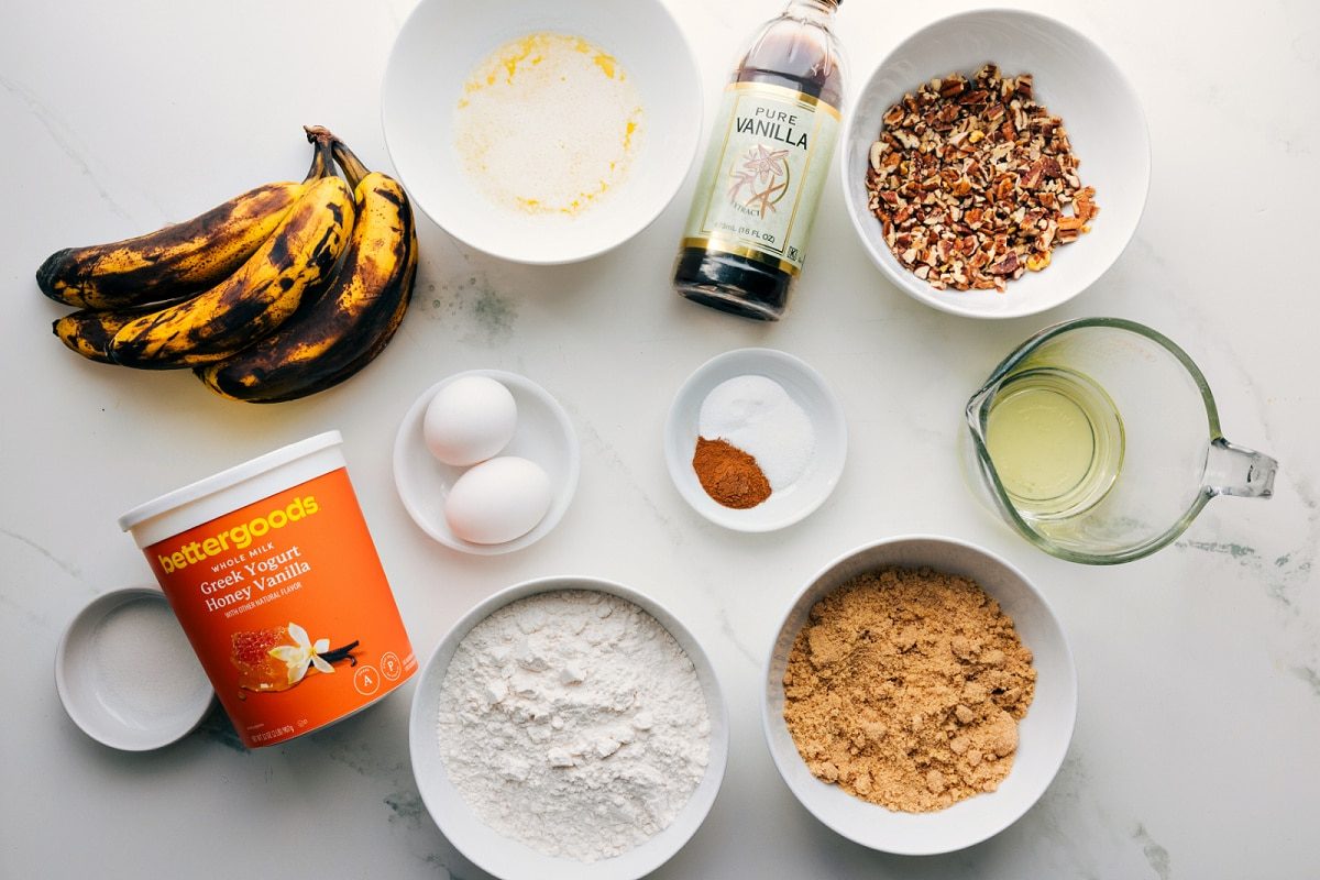 All the ingredients in this recipe prepped out for straightforward assembly including the bananas, yogurt, flour, sugar, oil, nuts, vanilla, spices, baking agents, eggs, and butter.