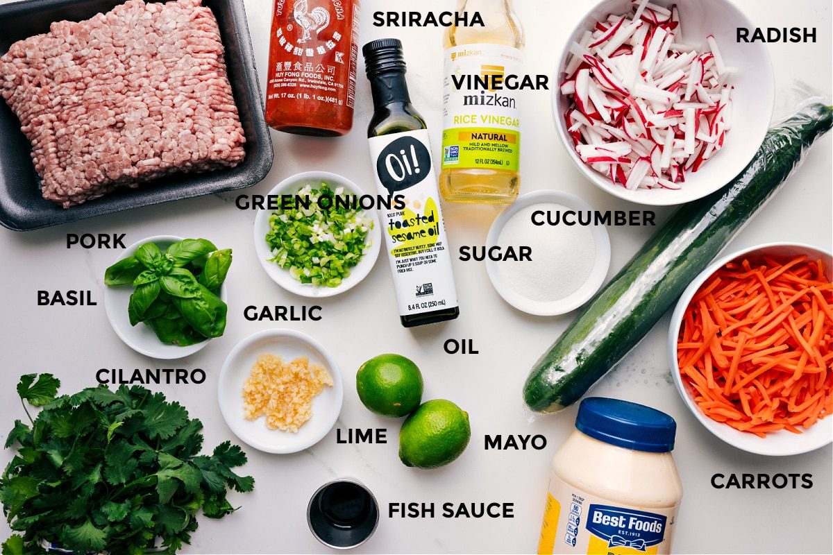 All the ingredients for this recipe prepped and ready for straightforward assembly, including pork, sriracha, vinegar, oil, green onions, basil, cilantro, garlic, lime, mayo, fish sauce, carrots, cucumber, sugar, radishes, and vinegar.