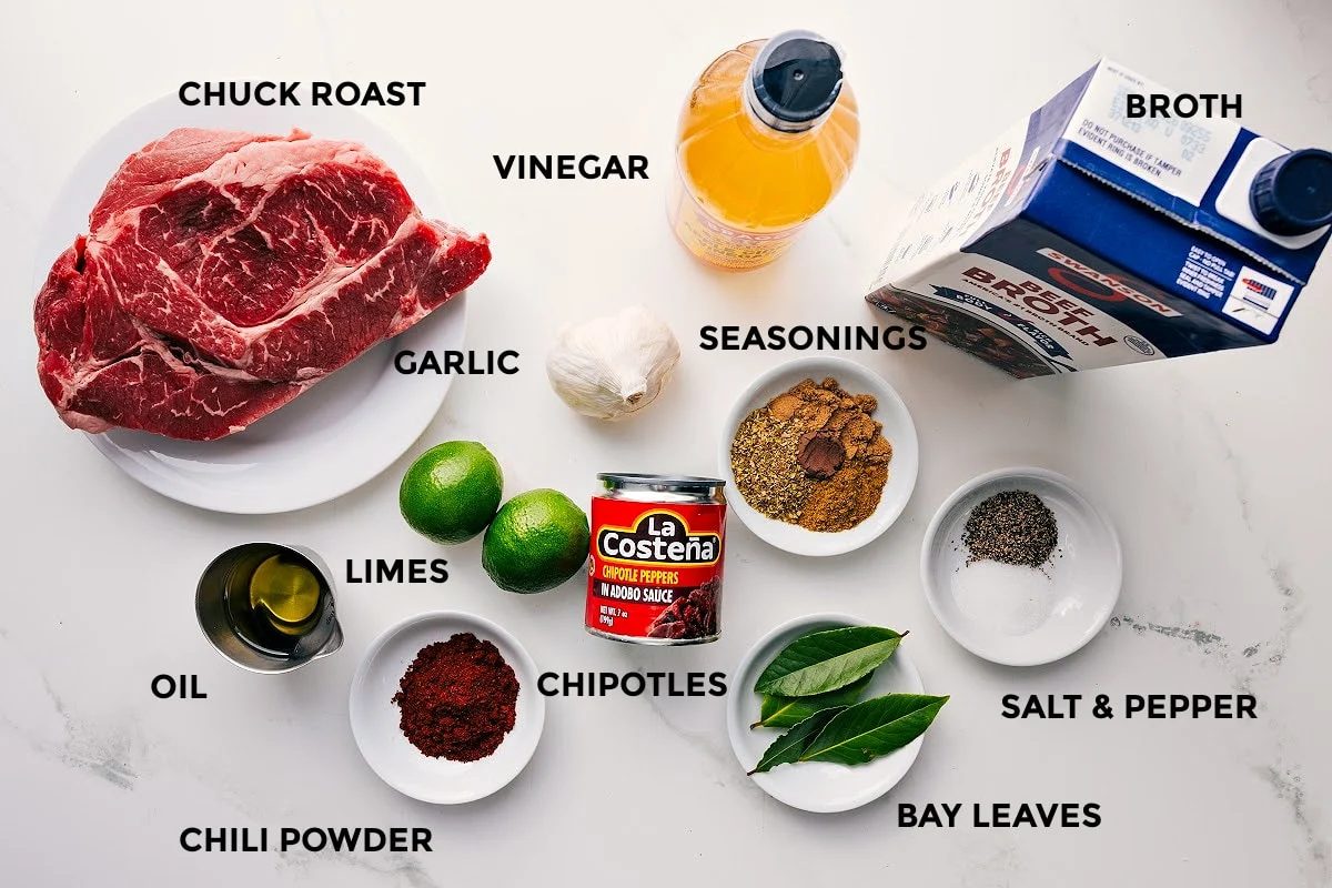All the ingredients in this recipe prepped out for straightforward assembly including the chuck roast, oil, seasonings, limes, chipotles, bay leaves, broth, and vinegar.