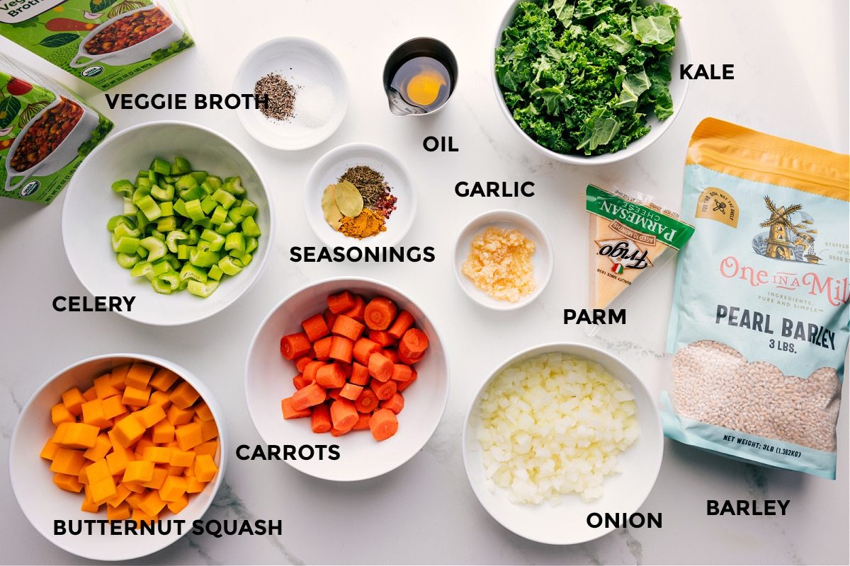 All the ingredients in this recipe prepped out for straightforward assembly including the broth, celery, butternut squash, carrots, seasonings, oil, garlic, onion, parmesan, kale, and barley.