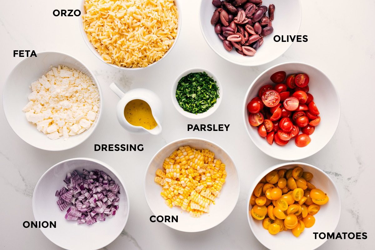 All the ingredients in this recipe prepped out for straightforward assembly including the feta, onion, dressing, corn, tomatoes, olives, parsley, and orzo.