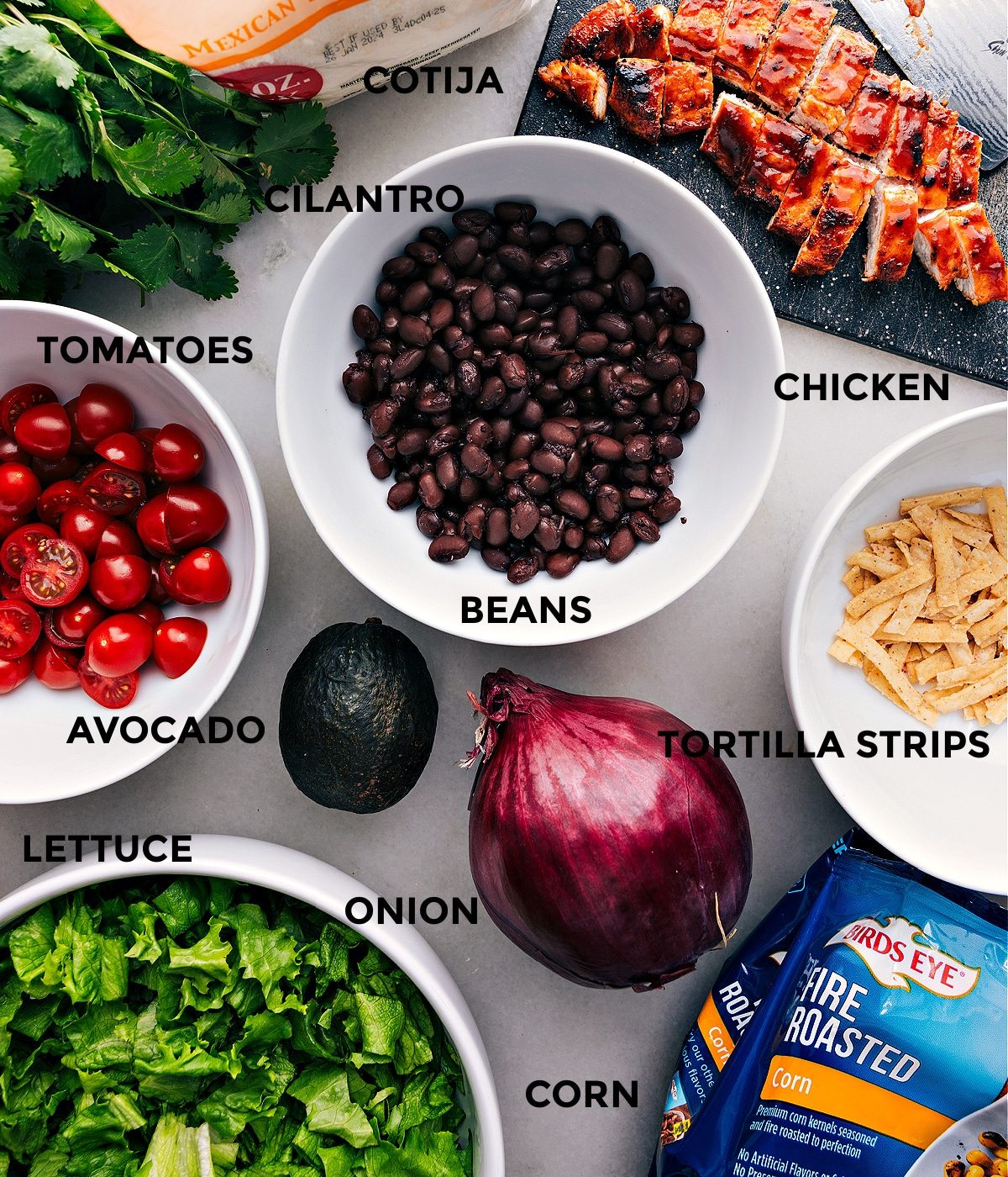 All the ingredients assembled, including cooked chicken, beans, tomatoes, cotija cheese, avocado, lettuce, onion, corn, and tortilla strips.