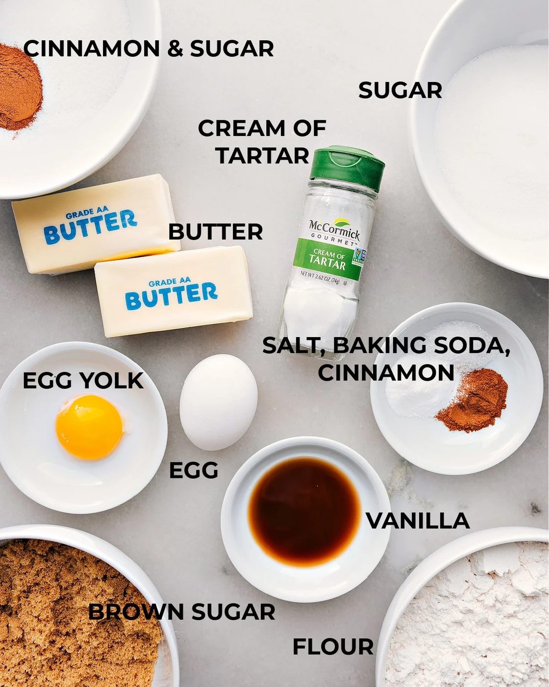 Various ingredients for making the snickerdoodle recipe arranged on a kitchen counter, including flour, sugar, eggs, and cinnamon.