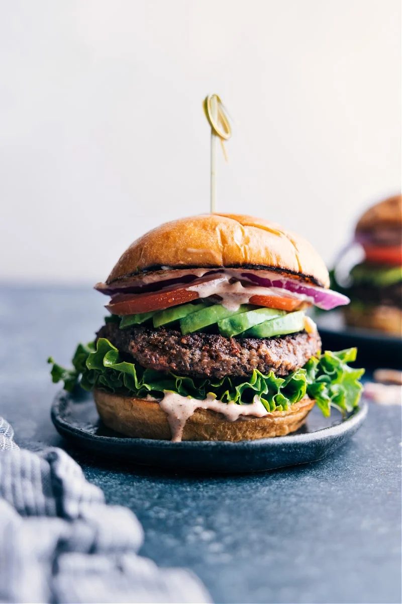 Plate with a satisfying black bean burger, topped with a colorful array of fresh vegetables and flavorful sauce dripping down.