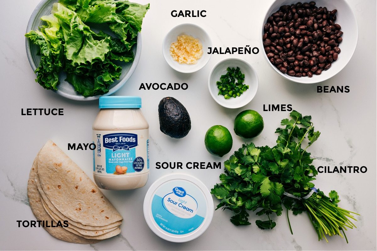 All the ingredients for this recipe prepped and ready for straightforward assembly, including lettuce, avocado, limes, beans, sour cream, mayo, cilantro, and tortillas.
