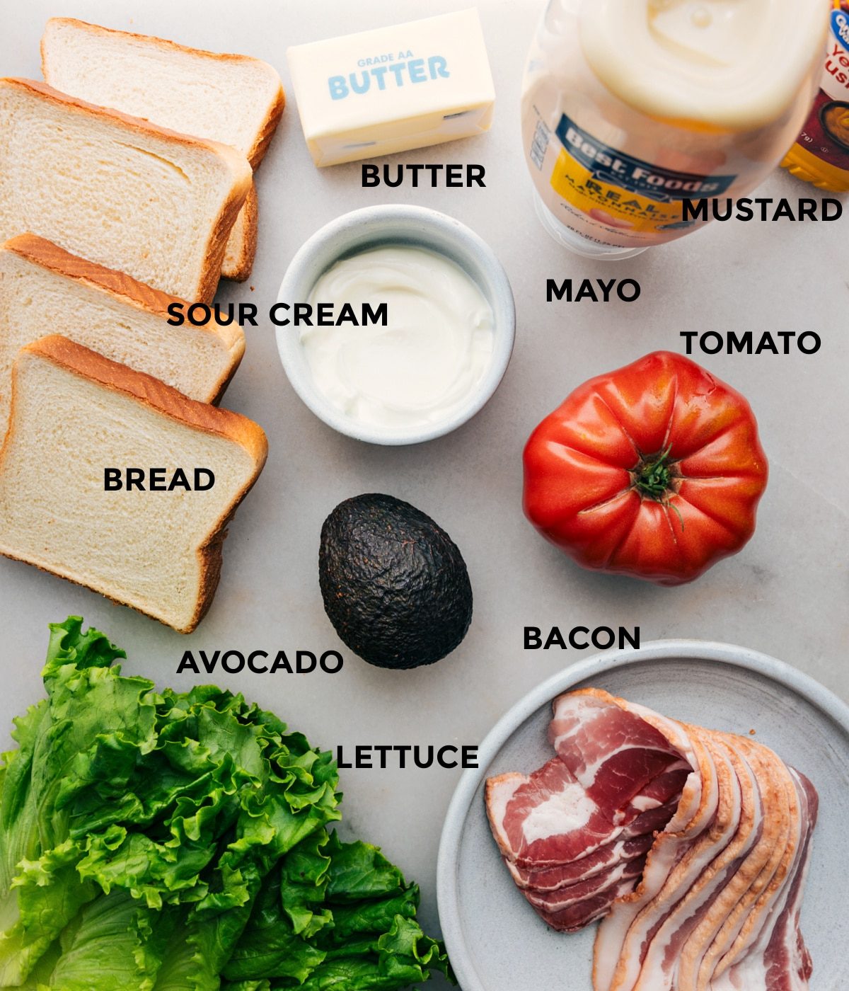 All the ingredients in this recipe including bread, sour cream, butter, mustard, mayo, tomato, bacon, avocado, and lettuce prepped out for straightforward assembly.