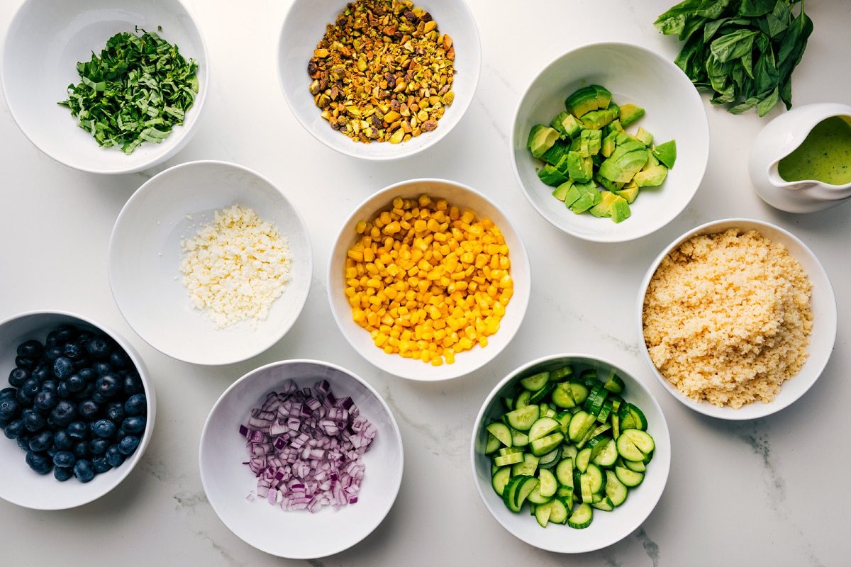 Ingredients for blueberry salad prepped for assembly with mint, blueberries, feta, red onion, pistachios, corn, cucumber, bulgur, avocado, and dressing.