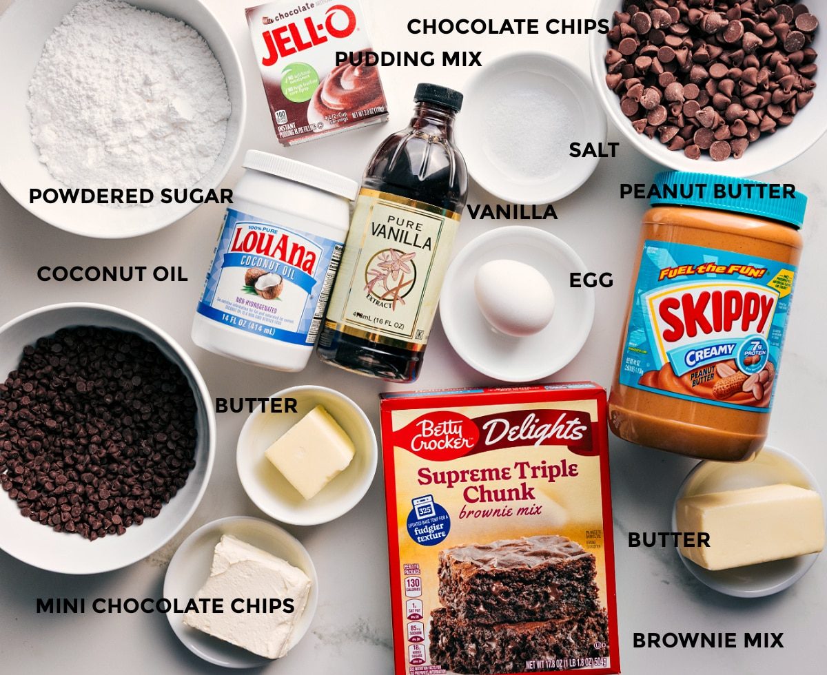 All the ingredients in this recipe prepped out for straightforward assembly including the powdered sugar, coconut oil, chocolate chips, butter, vanilla, pudding mix, peanut butter, egg, and brownie mix.