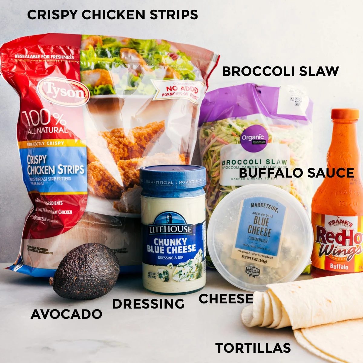 All the ingredients in this recipe, including crispy chicken strips, blue cheese dressing, sauce, broccoli slaw, tortillas, cheese, and avocado, are prepped and ready for straightforward assembly.