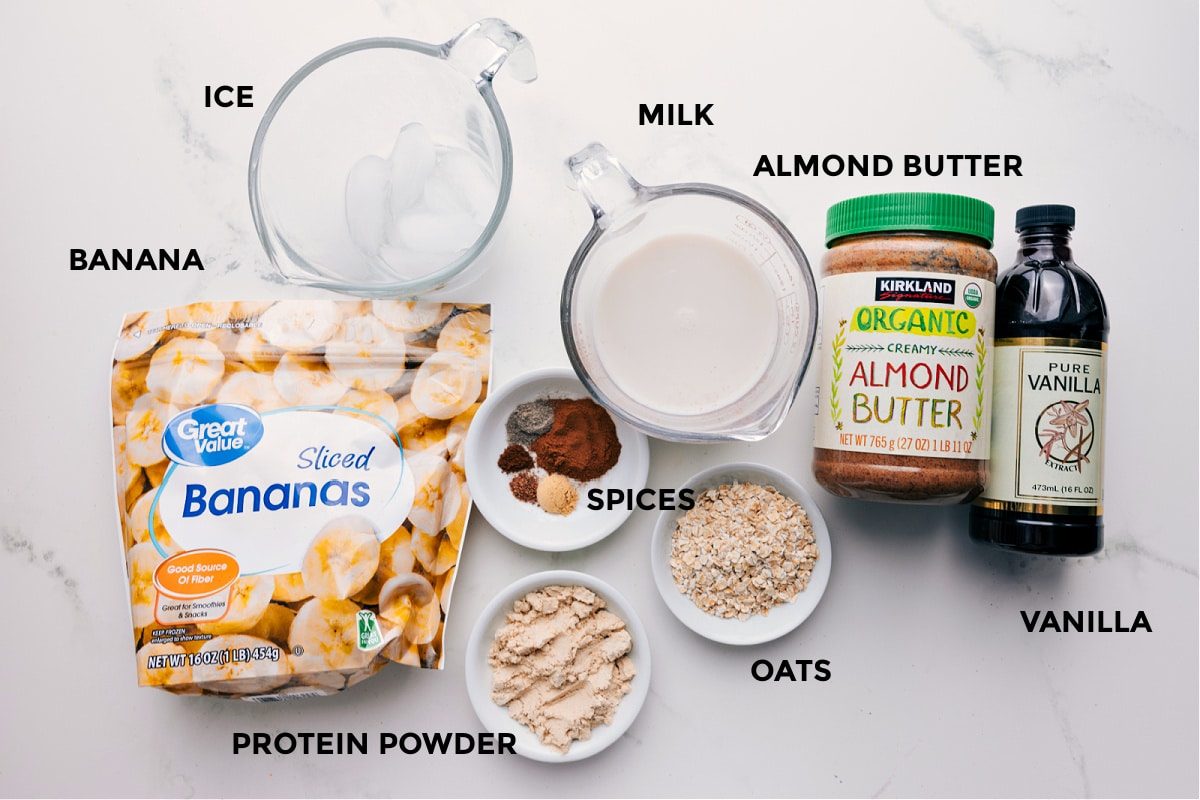 All the ingredients in this recipe prepped out for straightforward assembly including the banana, ice, spices, protein powder, oats, milk, almond butter, and vanilla.