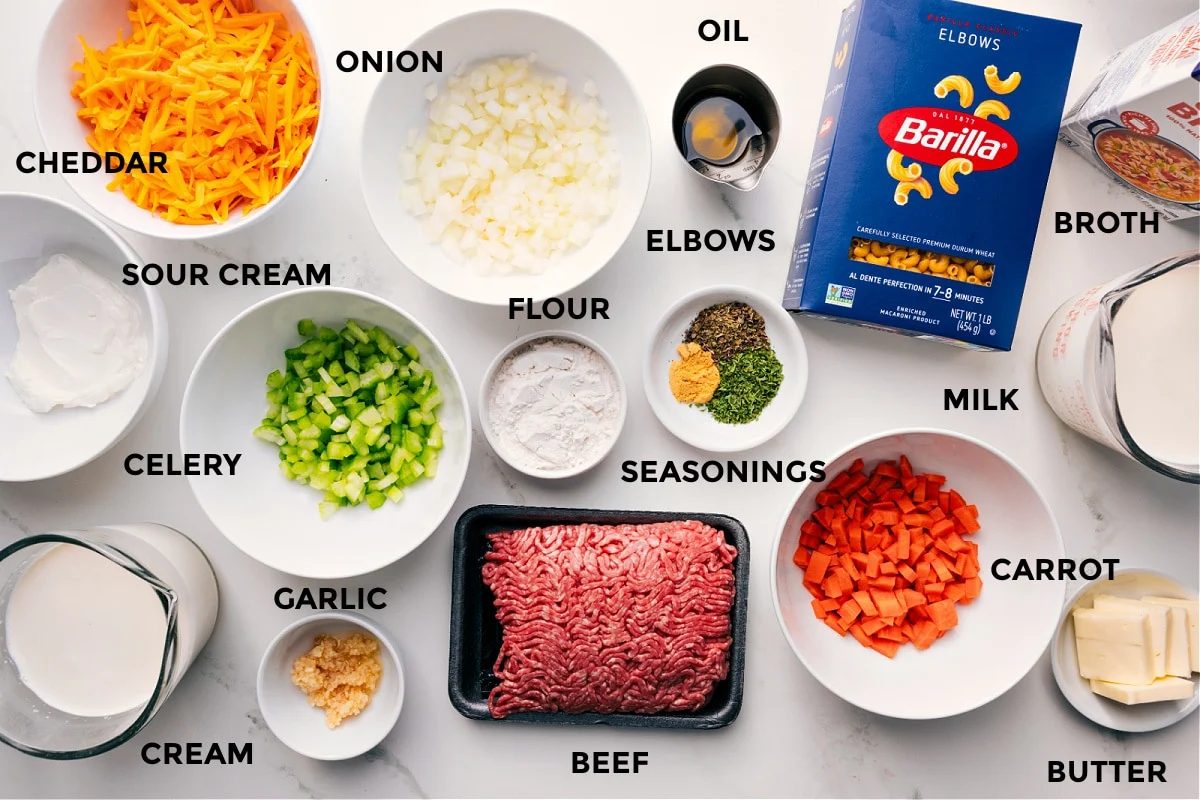 All the ingredients for this recipe prepped and ready for straightforward assembly, including sour cream, cream, cheddar, onion, celery, garlic, flour, ground beef, seasonings, oil, carrot, milk, butter, pasta, and broth.