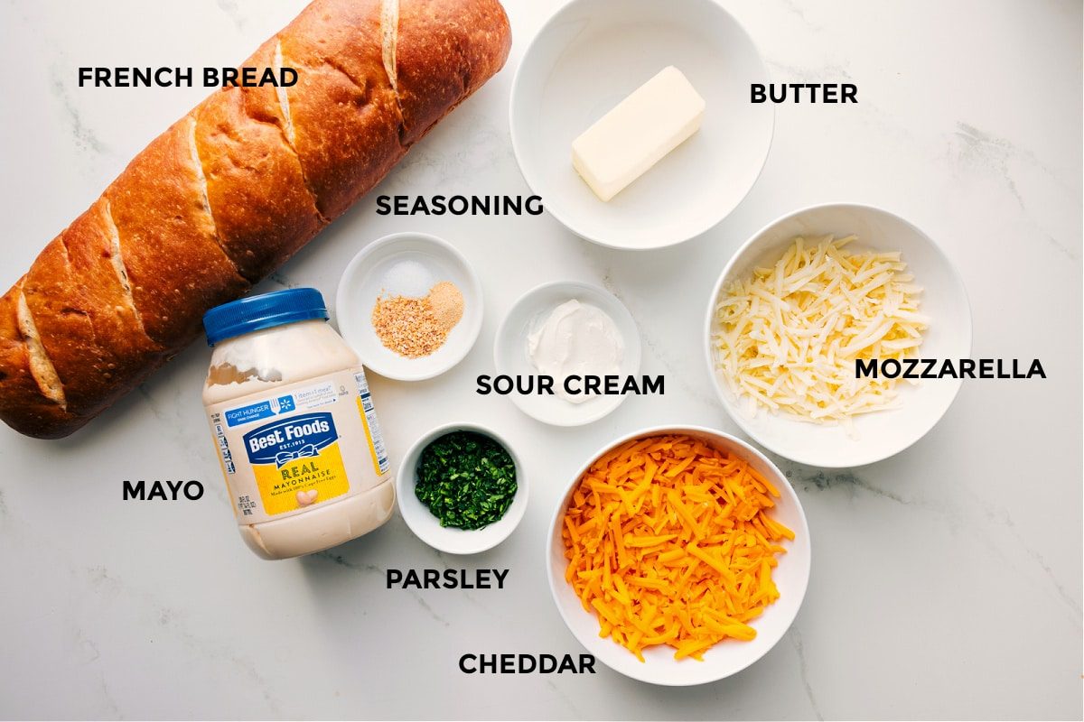All the ingredients prepped and ready to assemble, including French bread, mayo, seasonings, parsley, sour cream, butter, mozzarella, and cheddar.