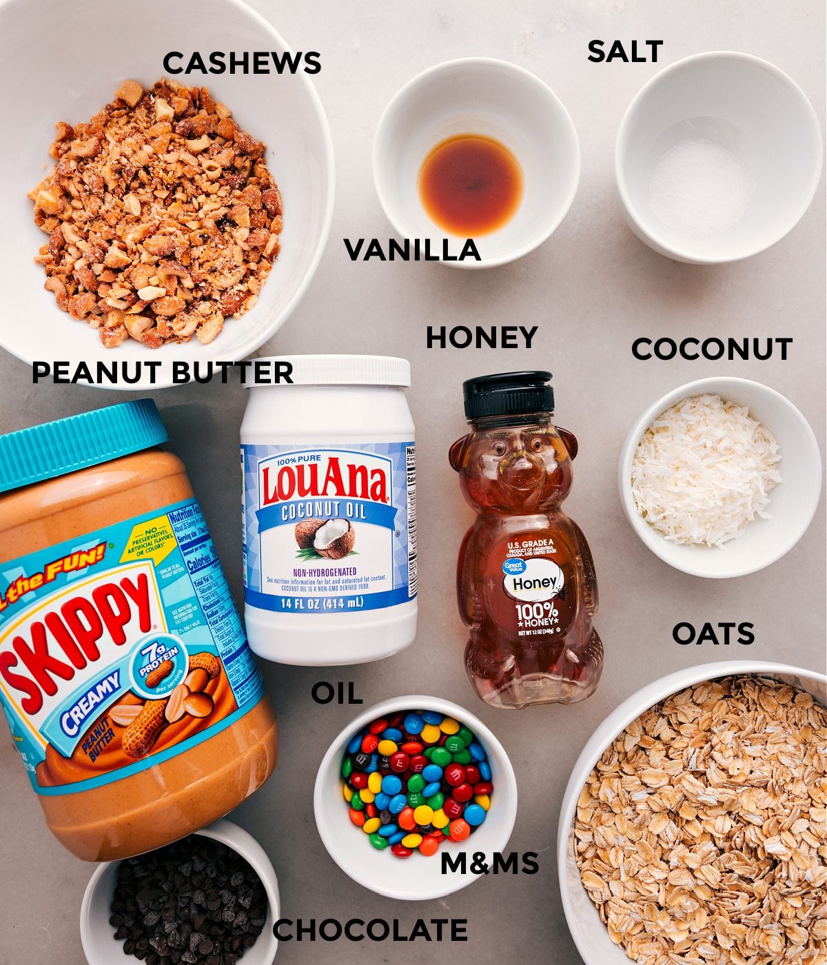 All the ingredients for this recipe are prepped and ready for straightforward assembly, including cashews, vanilla, salt, coconut, oats, honey, oil, peanut butter, M&Ms, and chocolate.