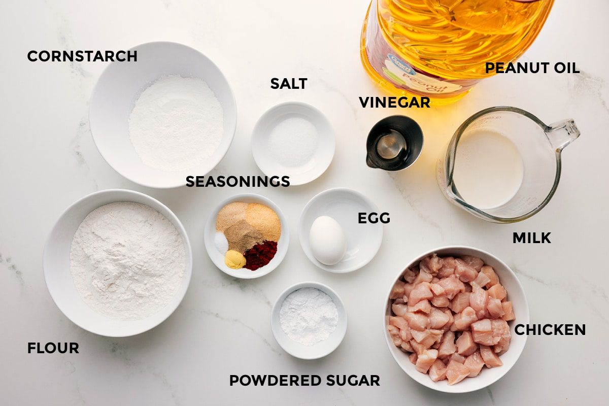 All the ingredients in this recipe prepped out for straightforward assembly including the cornstarch, flour, seasonings, salt, powdered sugar, egg, vinegar, peanut oil, milk, and chicken.