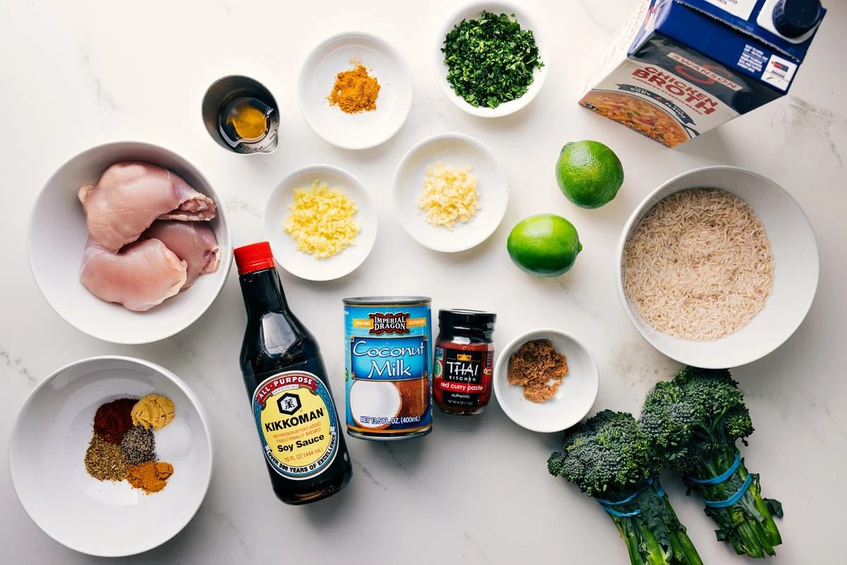 All the ingredients in this recipe prepped out for straightforward assembly including the chicken thighs, seasonings, garlic, ginger, soy sauce, coconut milk, curry paste, broccoli, rice, limes, herbs, and broth.
