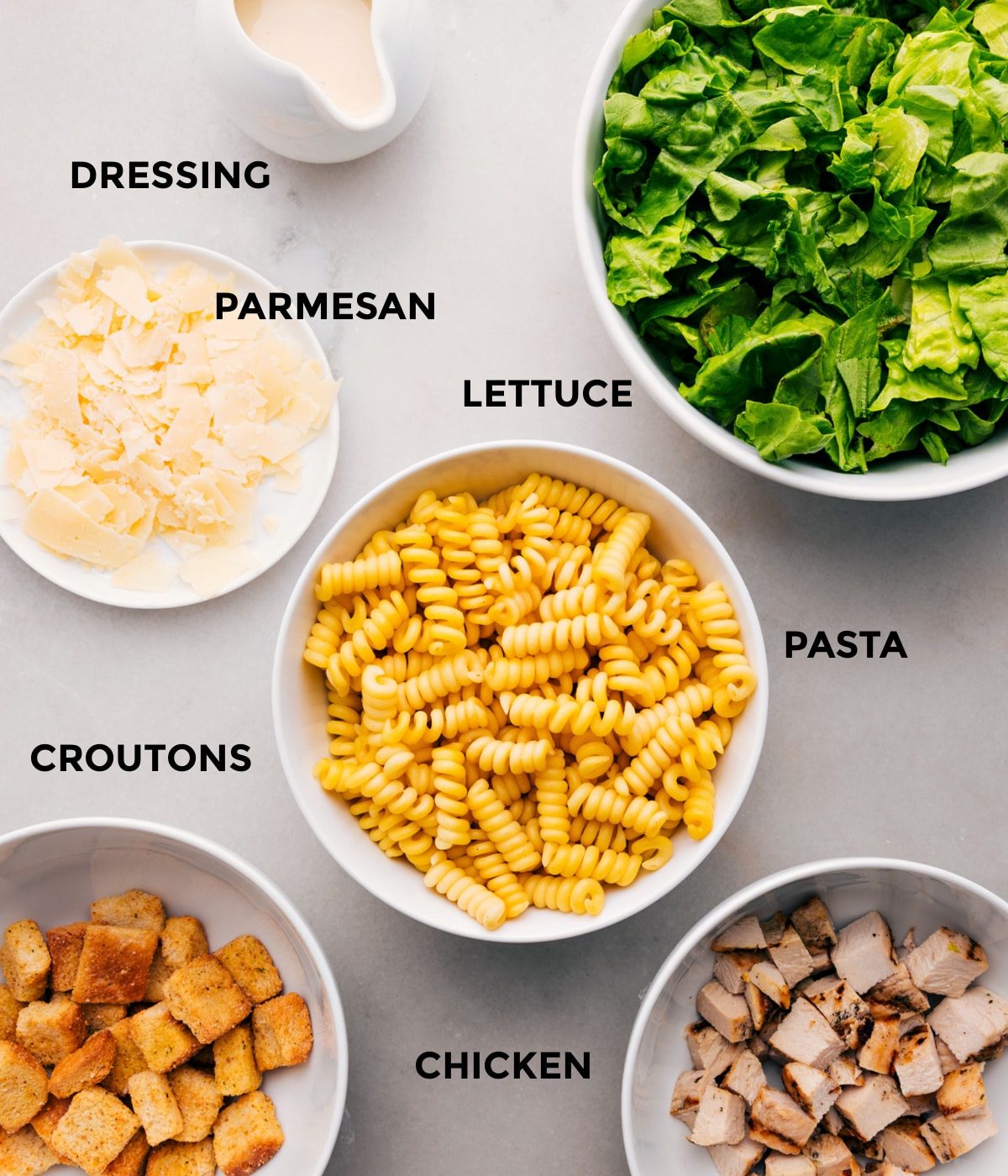 All the ingredients for this recipe prepped and ready for straightforward assembly, including the dressing, parmesan, lettuce, pasta, chicken, and croutons.
