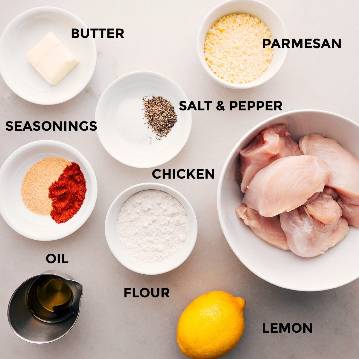 All the ingredients for this recipe are prepped and ready for straightforward assembly: parmesan, seasonings, chicken, flour, oil, and a lemon.