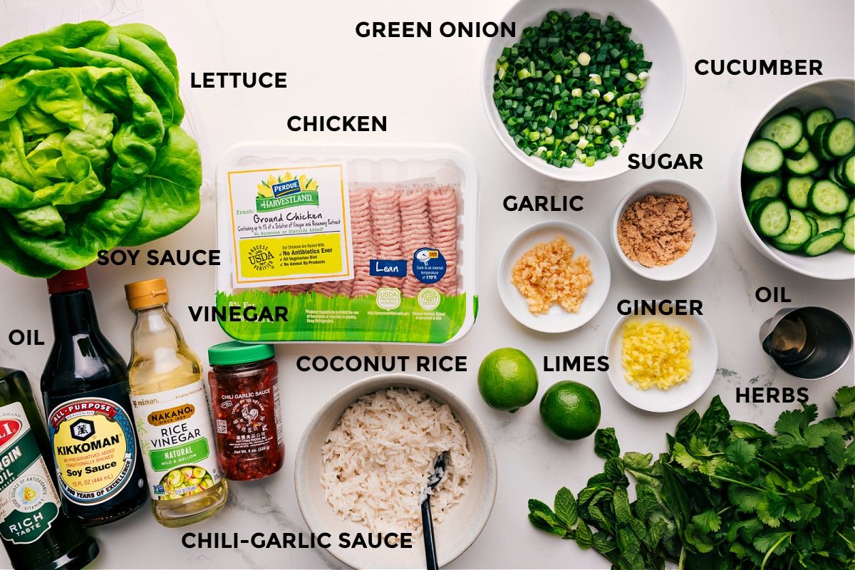 All the ingredients in this recipe prepped out for straightforward assembly including the lettuce, oil, soy sauce, vinegar, chili-garlic sauce, coconut rice, chicken, green onions, limes, garlic, ginger, sugar, cucumbers, and additional herbs.