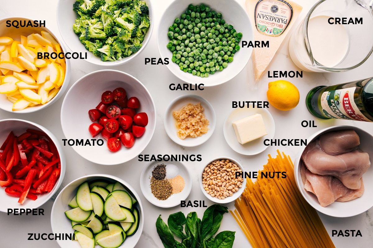 All the ingredients in this recipe prepped out for straightforward assembly including the squash, pepper, broccoli, zucchini, tomato, peas, garlic, seasonings, pine nuts, pasta, chicken, oil, lemon, cream, butter, parmesan, and lemon.