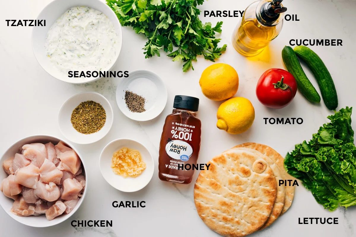 All the ingredients in this recipe prepped out for straightforward assembly including the tzatziki, seasonings, chicken, garlic, honey, pita, lettuce, tomato, cucumber, lemon, oil, parsley.