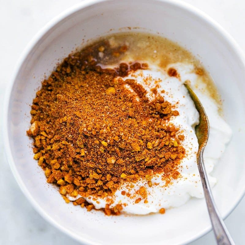 Marinade ingredients, including yogurt, lemon juice, garlic, ginger, and spices, being added to a bowl before being mixed together.
