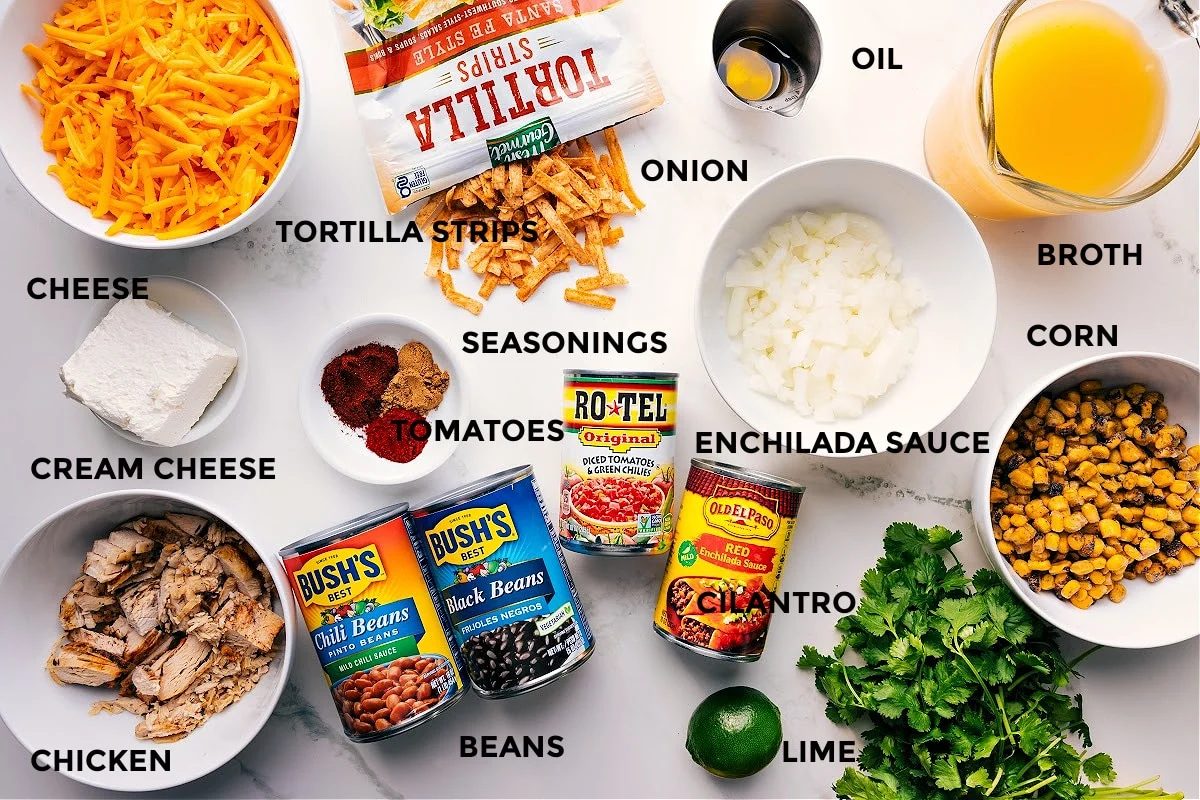 Chicken Tortilla Soup ingredients prepped and ready to assemble, including cheese, tortilla strips, cream cheese, chicken, beans, tomatoes, cilantro, onion, lime, enchilada sauce, corn, broth, oil, and seasonings.