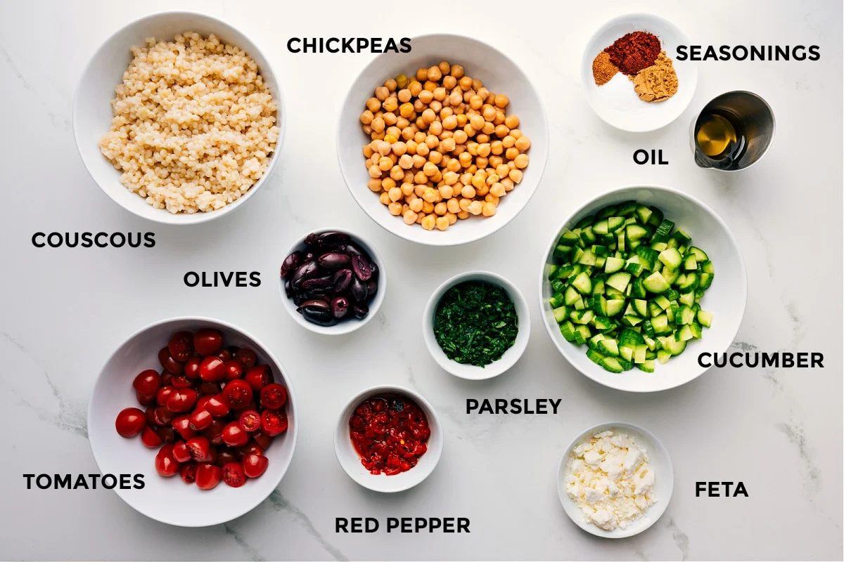 All the ingredients prepped for straightforward assembly, including chickpeas, couscous, seasonings, oil, cucumbers, parsley, feta, peppers, tomatoes, and olives.