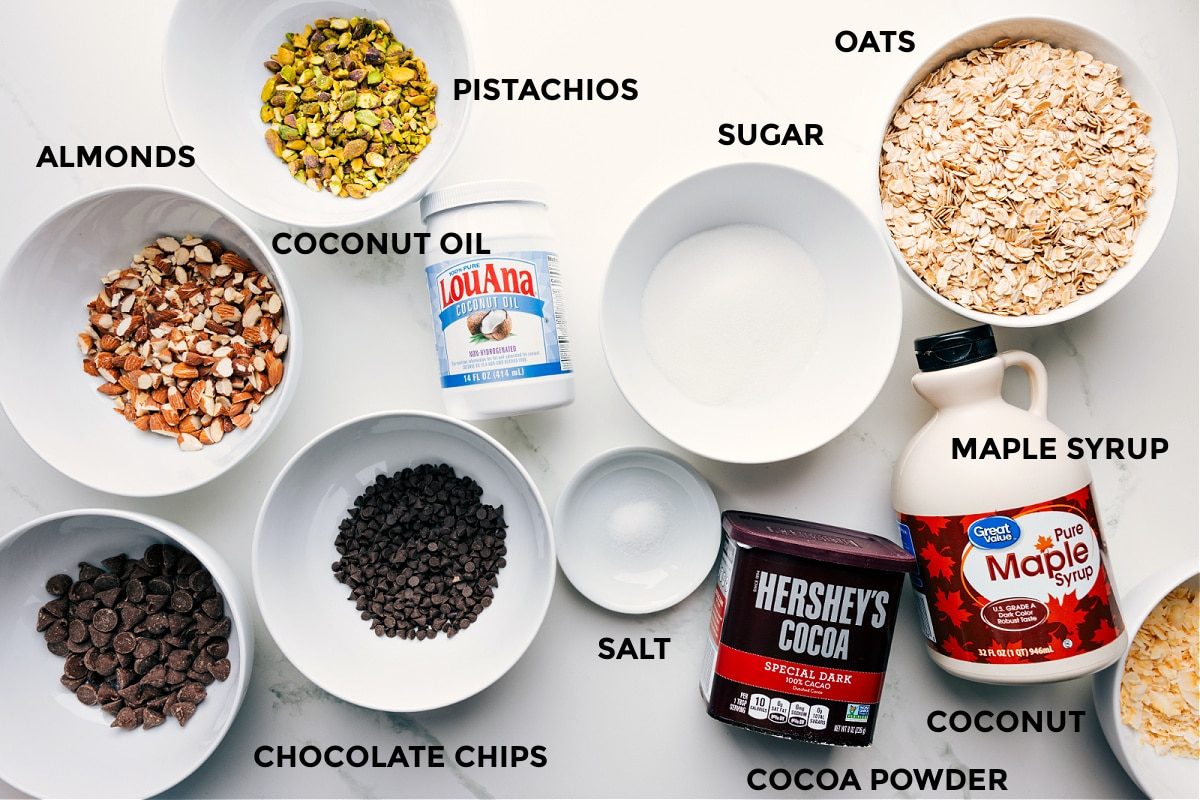 All the ingredients for this recipe laid out for straightforward assembly—oats, sugar, maple syrup, cocoa powder, salt, chocolate chips, coconut oil, pistachios, and almonds.