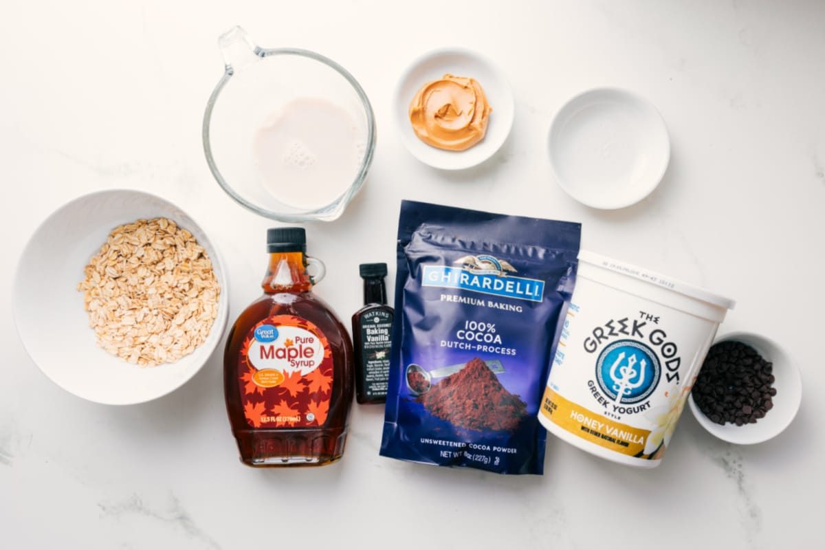All the ingredients in this recipe prepped out for straightforward assembly including the oats, milk, maple syrup, cocoa powder, vanilla, yogurt, chocolate chips, peanut butter, and salt.