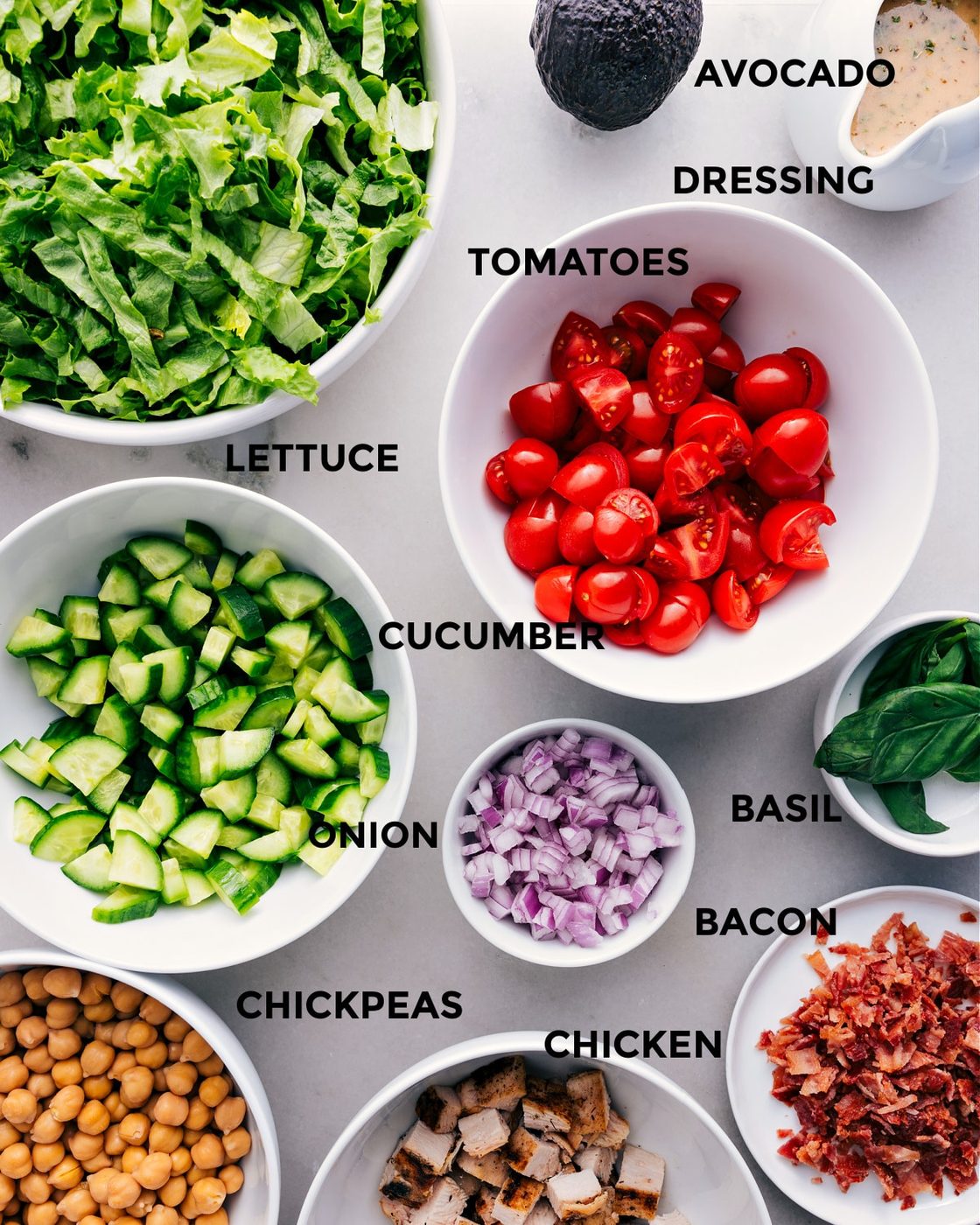 This recipe has everything prepped for straightforward assembly: tomatoes, avocado, dressing, lettuce, cucumbers, onions, basil, bacon, chicken, and chickpeas.