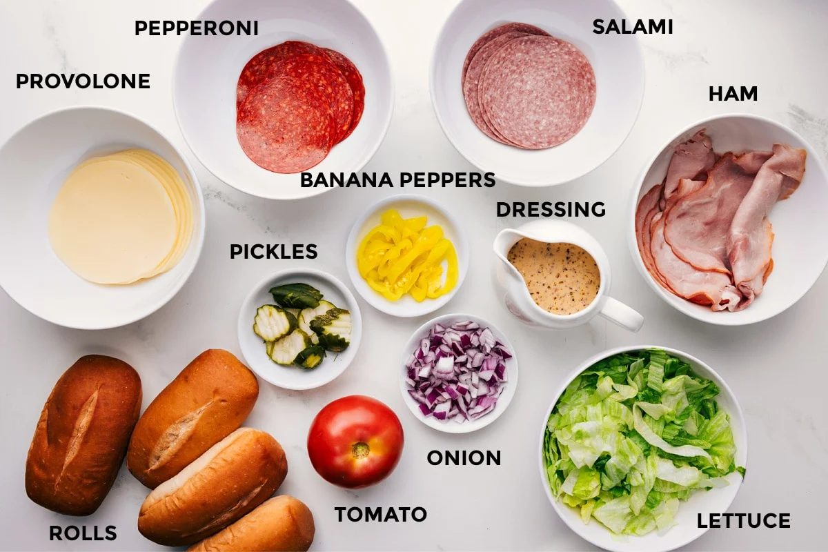 All the ingredients in this recipe prepped out for straightforward assembly including provolone, pepperoni, salami, ham, rolls, pickles, banana peppers, tomato, onion, dressing, and lettuce.