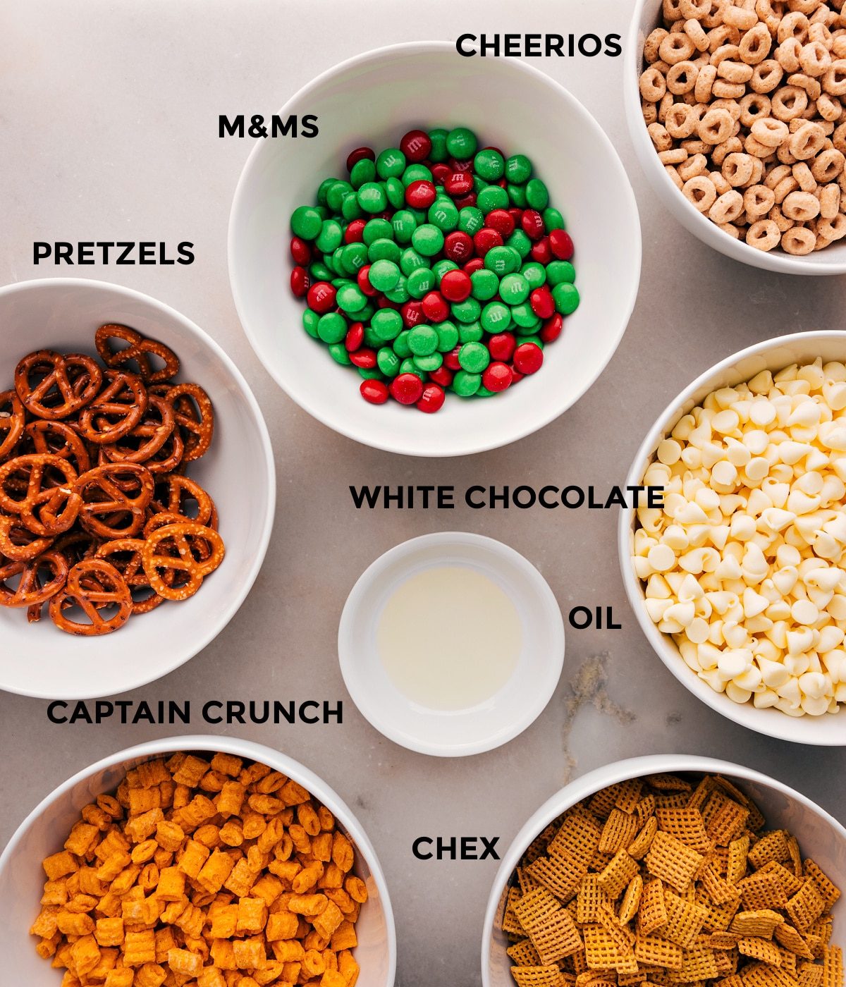 All the ingredients in this recipe are prepped for straightforward assembly: M&Ms, white chocolate, oil, Chex, Captain Crunch, and pretzels.
