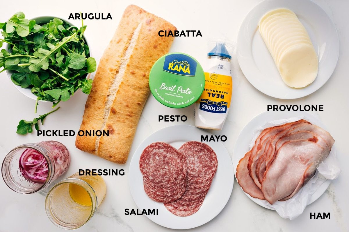 All the ingredients in this recipe prepped out for straightforward assembly including the arugula, pickled onions, dressing, ciabatta, pesto, mayo, salami, ham, and provolone.