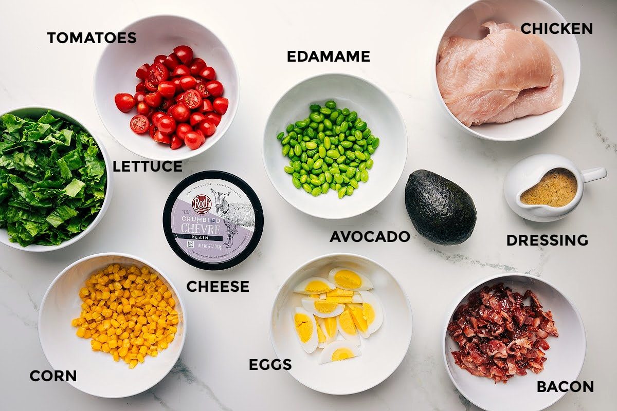 All the ingredients in this recipe prepped out for straightforward assembly including the lettuce, tomatoes, corn, cheese, edamame, eggs, avocado, dressing, bacon, and chicken.