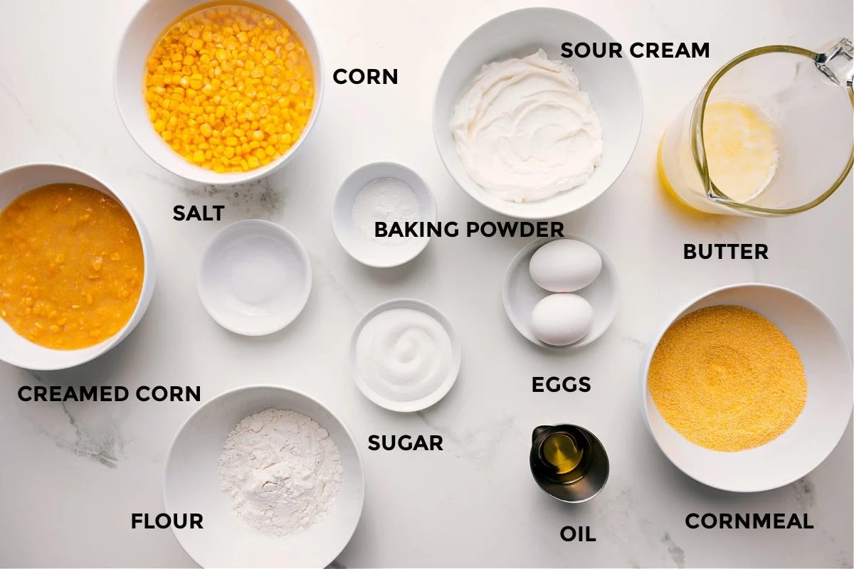 All the ingredients in this recipe prepped out for straightforward assembly including the creamed corn, corn, salt, flour, baking powder, sugar, sour cream, eggs, oil, cornmeal, and butter.