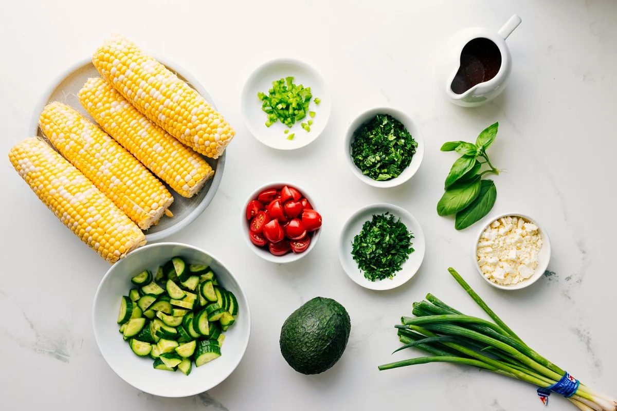 All the ingredients in this recipe prepped out for straightforward assembly including the corn, cucumbers, avocado, tomatoes, jalapeños, cilantro, parsley, basil, green onions, feta, and vinaigrette.