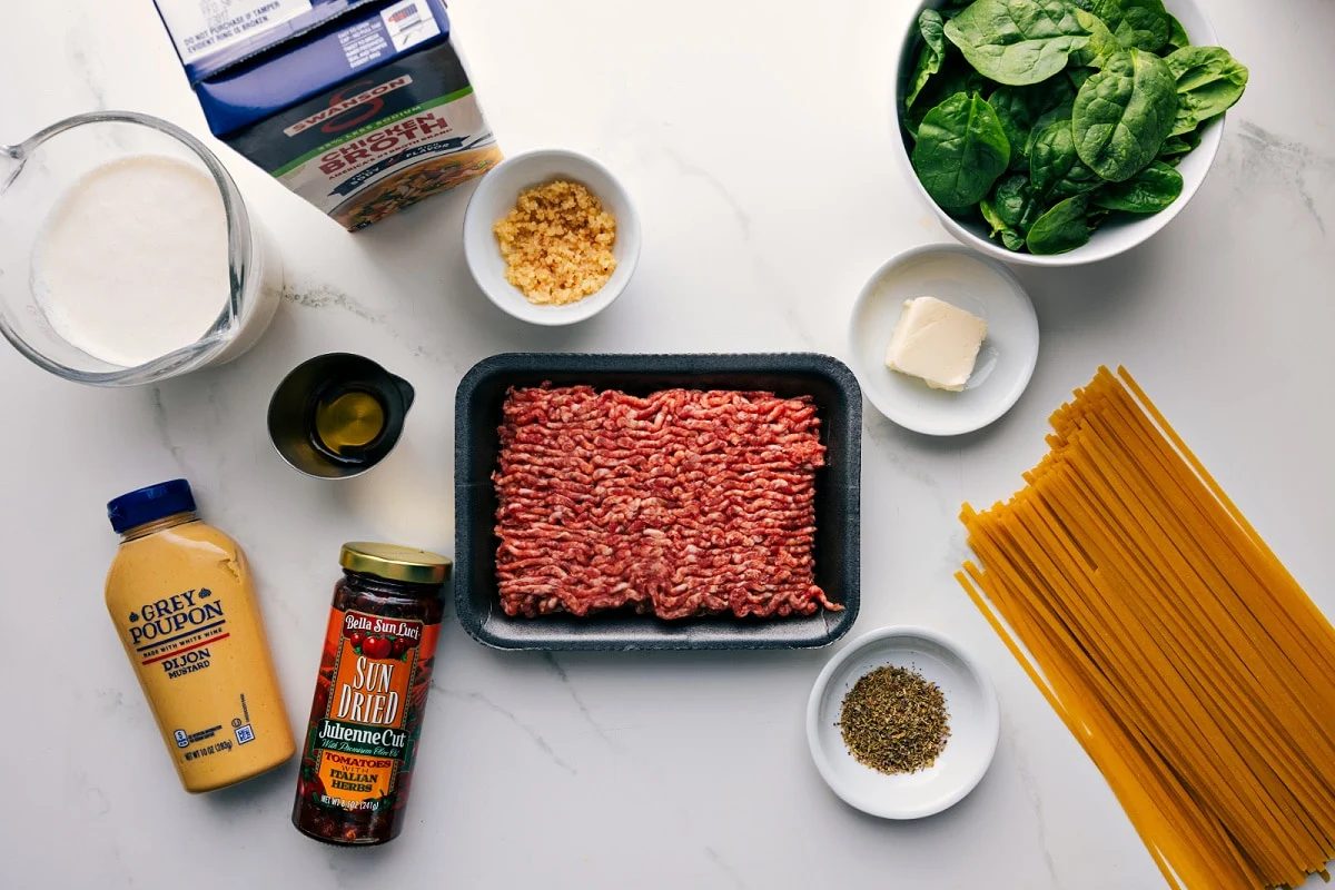 All the ingredients in this recipe prepped out for straightforward assembly including the cream, dijon, sun-dried tomatoes, broth, garlic, sausage, seasonings, spinach, butter, and pasta.
