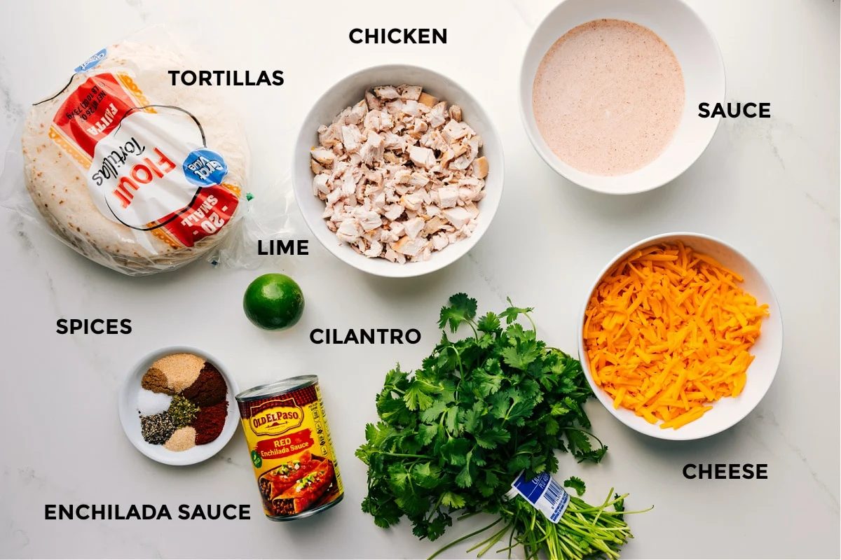 All the ingredients for this recipe prepped and ready for straightforward assembly, including chicken, sauce, cheese, cilantro, enchilada sauce, spices, tortillas, and limes.