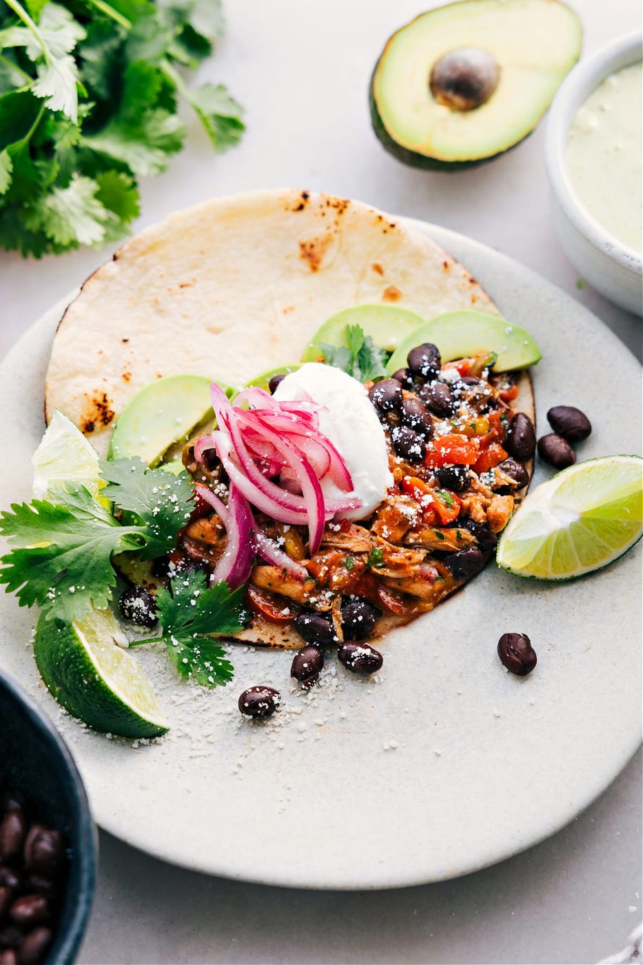 A slow cooker Chicken Fajita assembled in a tortilla with toppings like black beans, onions, sour cream, avocado, cheese, and limes.