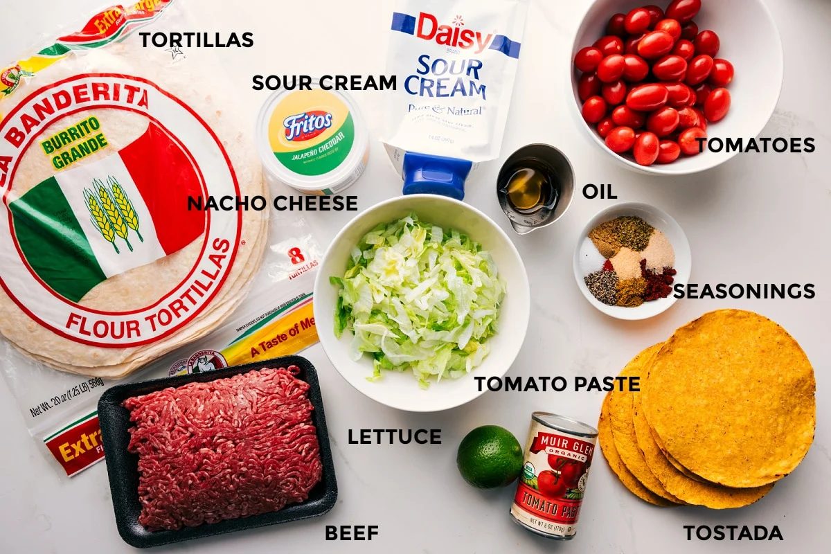 All the ingredients in this recipe prepped out for straightforward assembly including the tortillas, beef, lettuce, tomato paste, lime, seasonings, tomatoes, oil, sour cream, and nacho cheese.